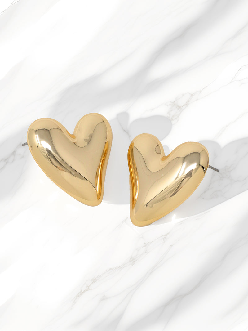 A pair of glossy gold puffy heart stud earrings with a high-shine finish, displayed on a white marble background. The earrings have a bold, statement design with a smooth, reflective surface.