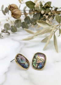 Gold-framed abalone shell earrings on a marble surface with green leaves.