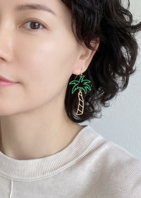 A pair of cutout palm tree drop earrings featuring bright green palm leaves and a gold-tone trunk, dangling from gold hook backings.