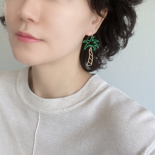 A woman wearing a pair of chic tropical palm tree earrings with dangling design, showcasing green epoxy-coated tops resembling tropical leaves.