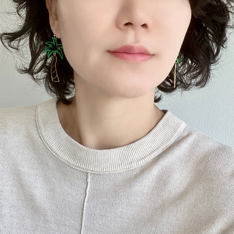 A woman wearing a pair of chic tropical palm tree earrings with dangling design, showcasing green epoxy-coated tops resembling tropical leaves.