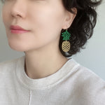 A woman wearing chic pineapple earrings with dangling design, showcasing green epoxy-coated tops resembling tropical leaves.