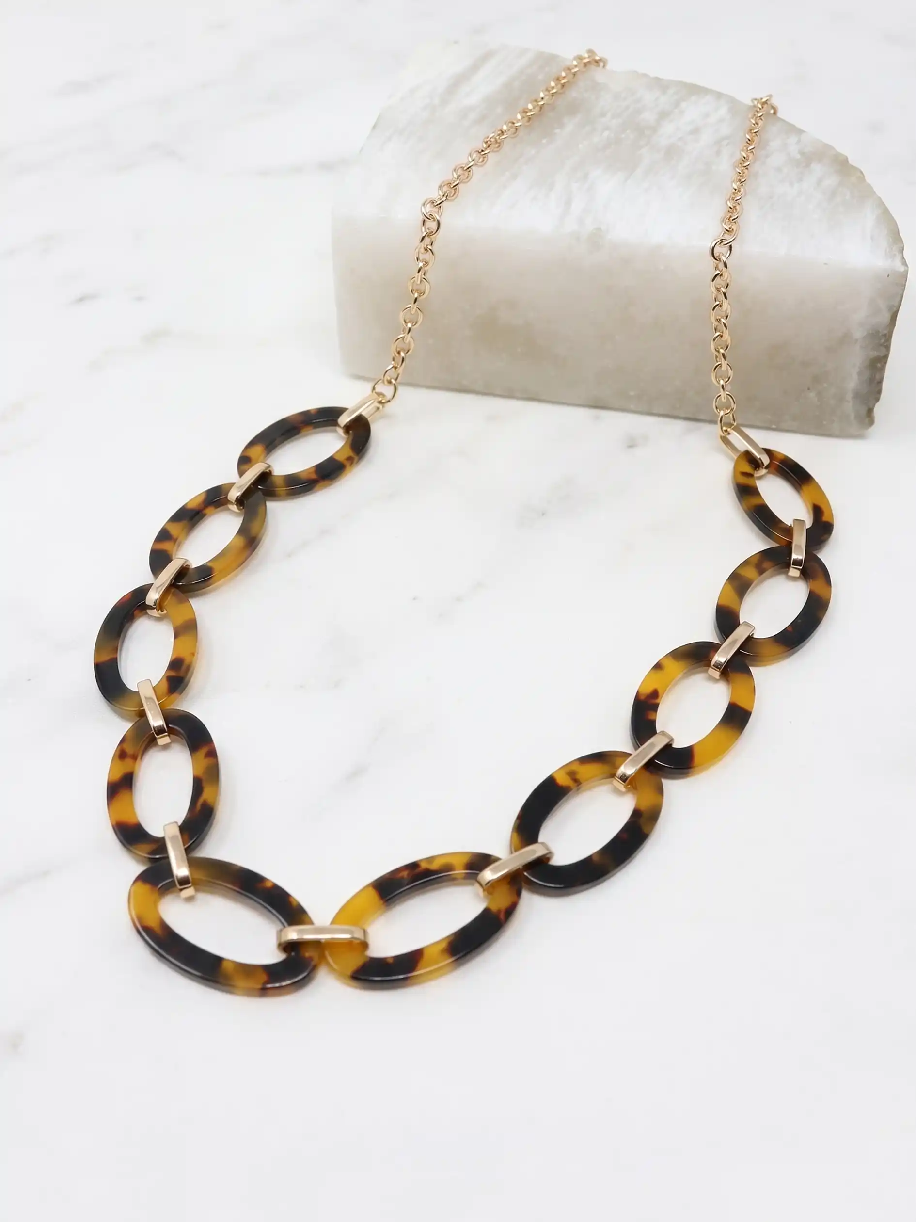 A gold-tone chain necklace featuring large, polished brown tortoise resin oval links, displayed on a white marble surface.