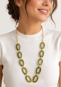 A close-up of a woman wearing a gold-tone chain necklace with large olive green oval links, paired with a white ribbed short-sleeve top.