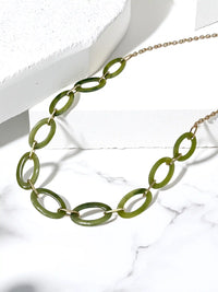 A gold-tone chain necklace featuring large, polished olive green oval links, displayed on a white marble surface with a white geometric block.