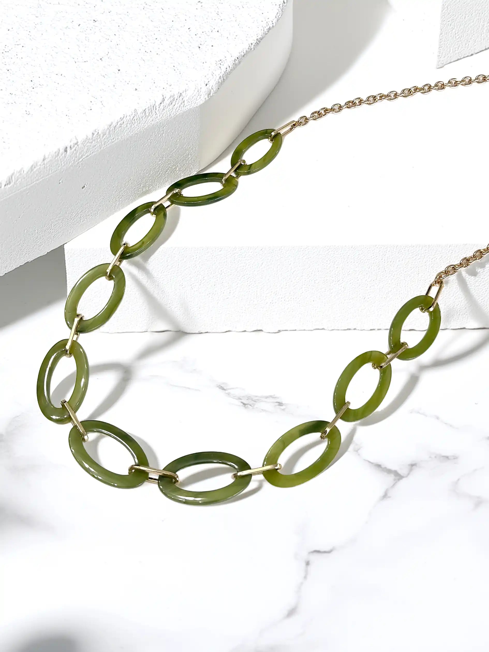 A gold-tone chain necklace featuring large, polished olive green oval links, displayed on a white marble surface with a white geometric block.