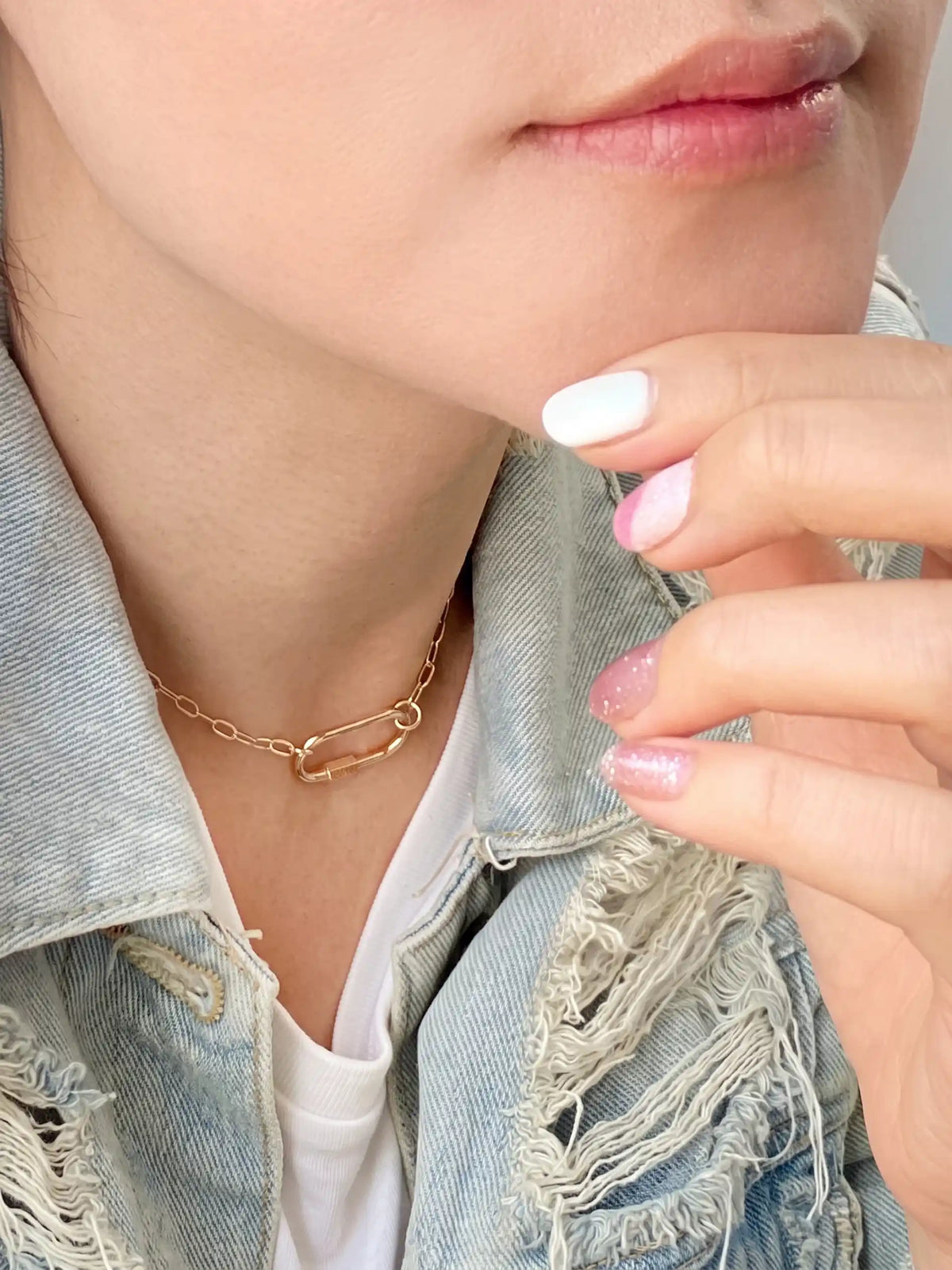 A close-up lifestyle shot of a model wearing a delicate gold chain necklace with a safety-pin style connector. The model is wearing a light-wash distressed denim jacket over a white t-shirt, with her hand resting near her chin showing white and pink manicured nails.