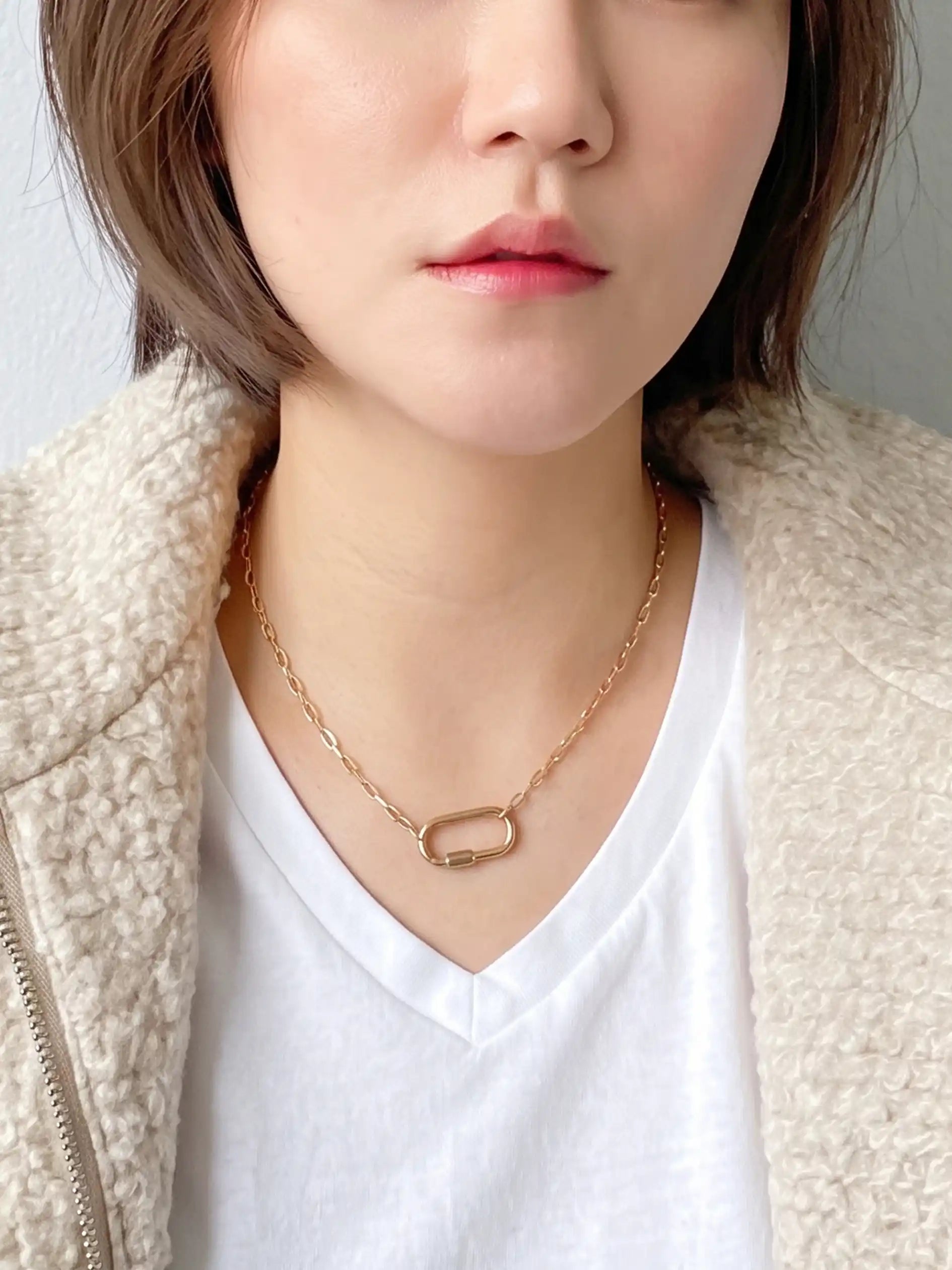 A close-up of a person wearing a delicate gold chain necklace with a small rectangular link pendant, styled with a white V-neck t-shirt and a textured cream fleece jacket.