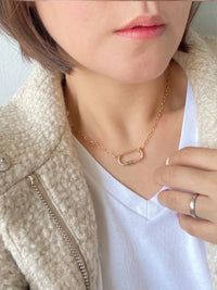 A close-up lifestyle shot of a model wearing a delicate rose gold chain necklace with a small open-link safety-pin style pendant. The model is wearing a white V-neck t-shirt and a textured cream-colored jacket.