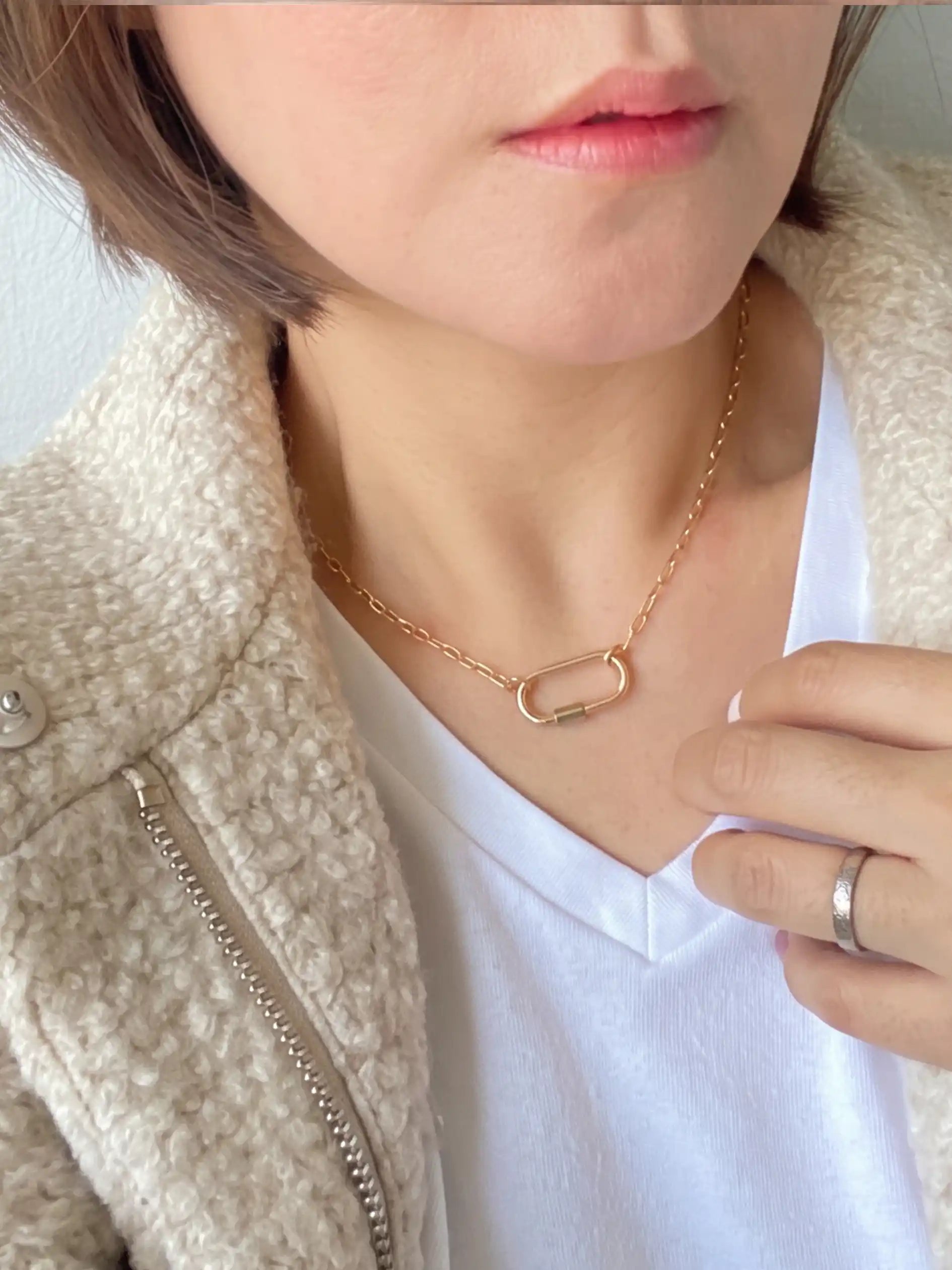 A close-up lifestyle shot of a model wearing a delicate rose gold chain necklace with a small open-link safety-pin style pendant. The model is wearing a white V-neck t-shirt and a textured cream-colored jacket.