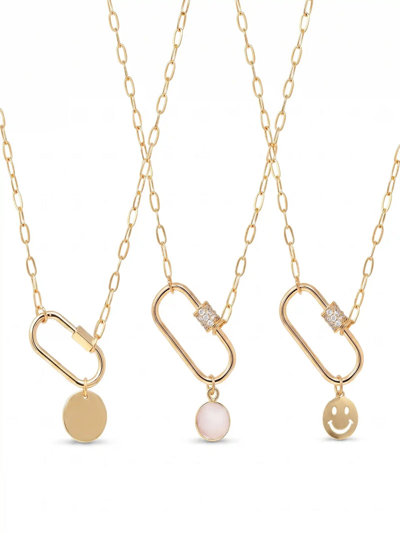 Three gold chain necklaces with elongated oval pendants are displayed against a white background. The necklaces feature different charms: a plain round disc, a pale pink oval stone, and a gold smiley face.