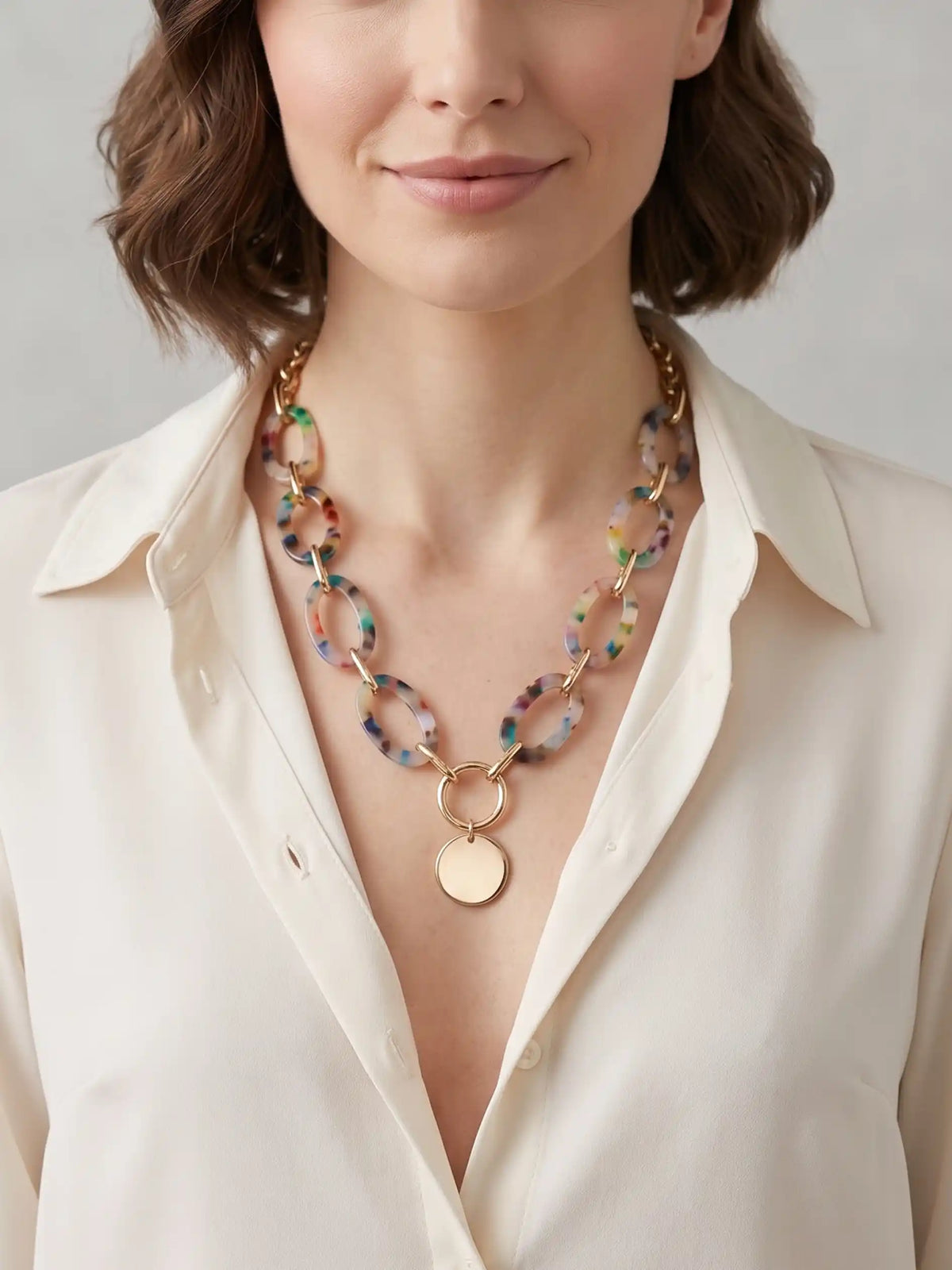 A woman wearing an ivory open-collar shirt showcasing a chunky multi-color resin oval link statement necklace with a gold-tone center ring and a round flat gold pendant.