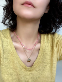 A close-up of a person wearing layered necklaces, including a pink beaded chain, a delicate gold chain, and a gold pendant necklace, paired with a yellow top.
