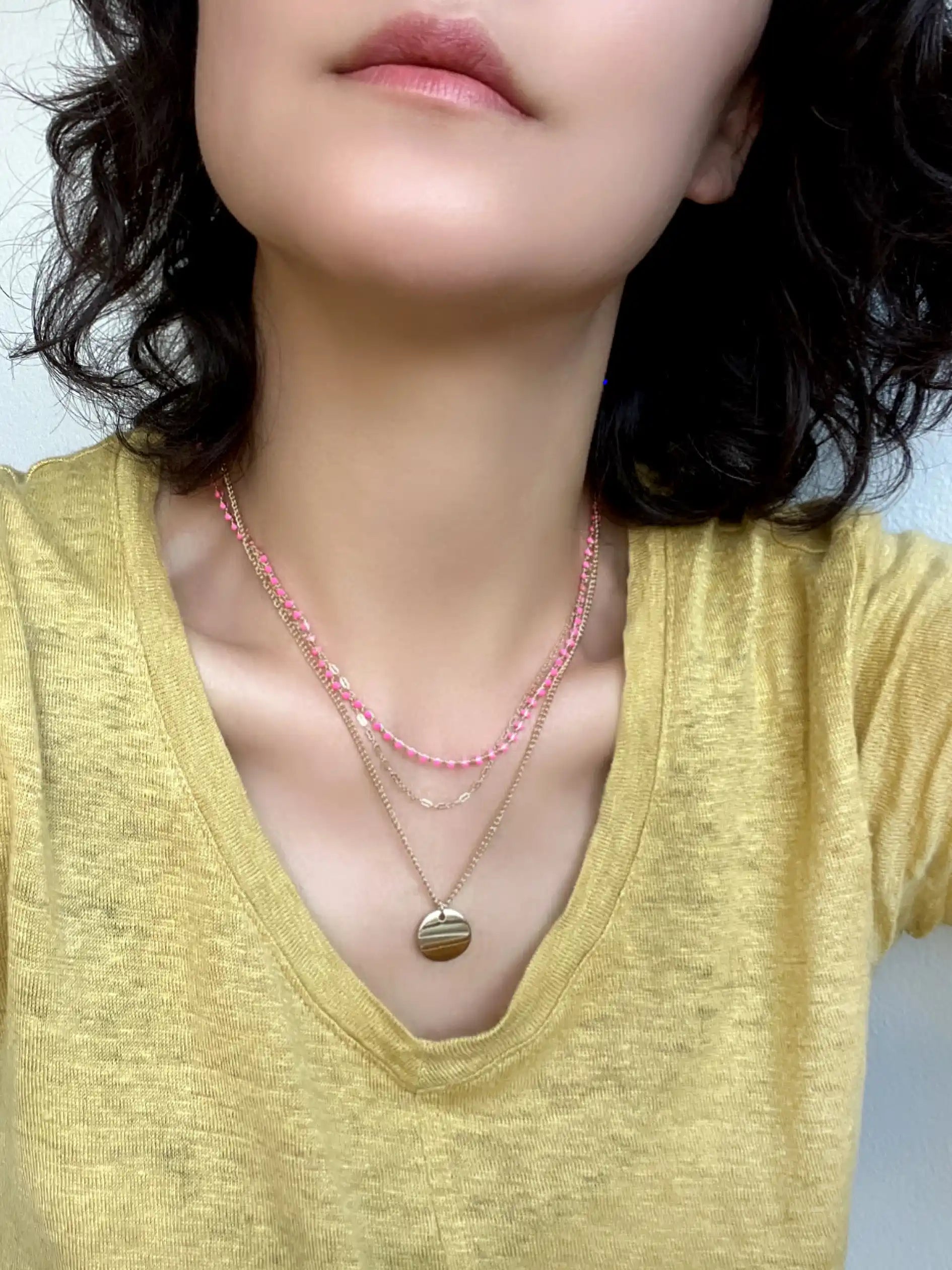 A close-up of a person wearing layered necklaces, including a pink beaded chain, a delicate gold chain, and a gold pendant necklace, paired with a yellow top.