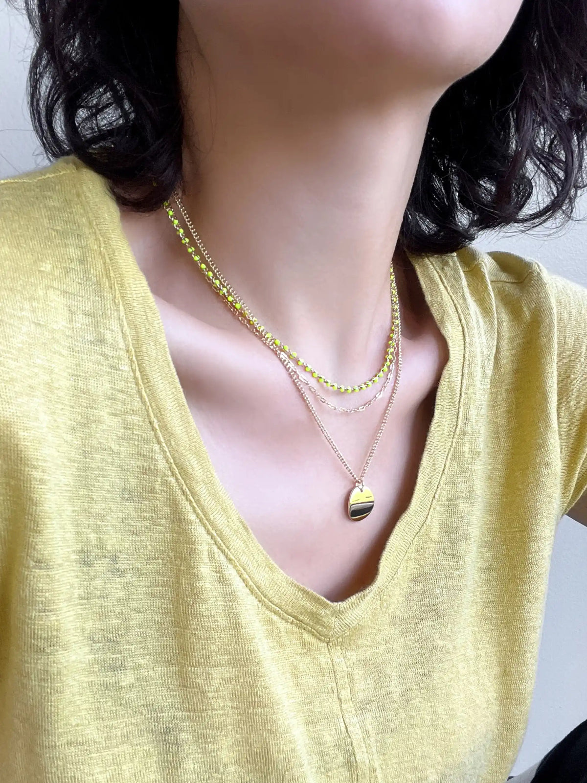 A close-up of a person wearing layered necklaces, including a gold chain with a small rainbow-striped heart pendant and a yellow beaded chain, paired with a yellow-green V-neck top.