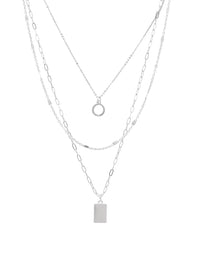 A layered silver necklace set featuring three delicate chains: a fine chain with a small circular ring pendant, a chain with a rectangular bar pendant, and a chain with elongated oval links, all on a plain white background.