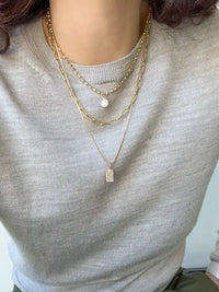 A close-up of a person wearing a layered gold necklace set with a small rectangular pendant and a round charm, styled over a light gray knit top.