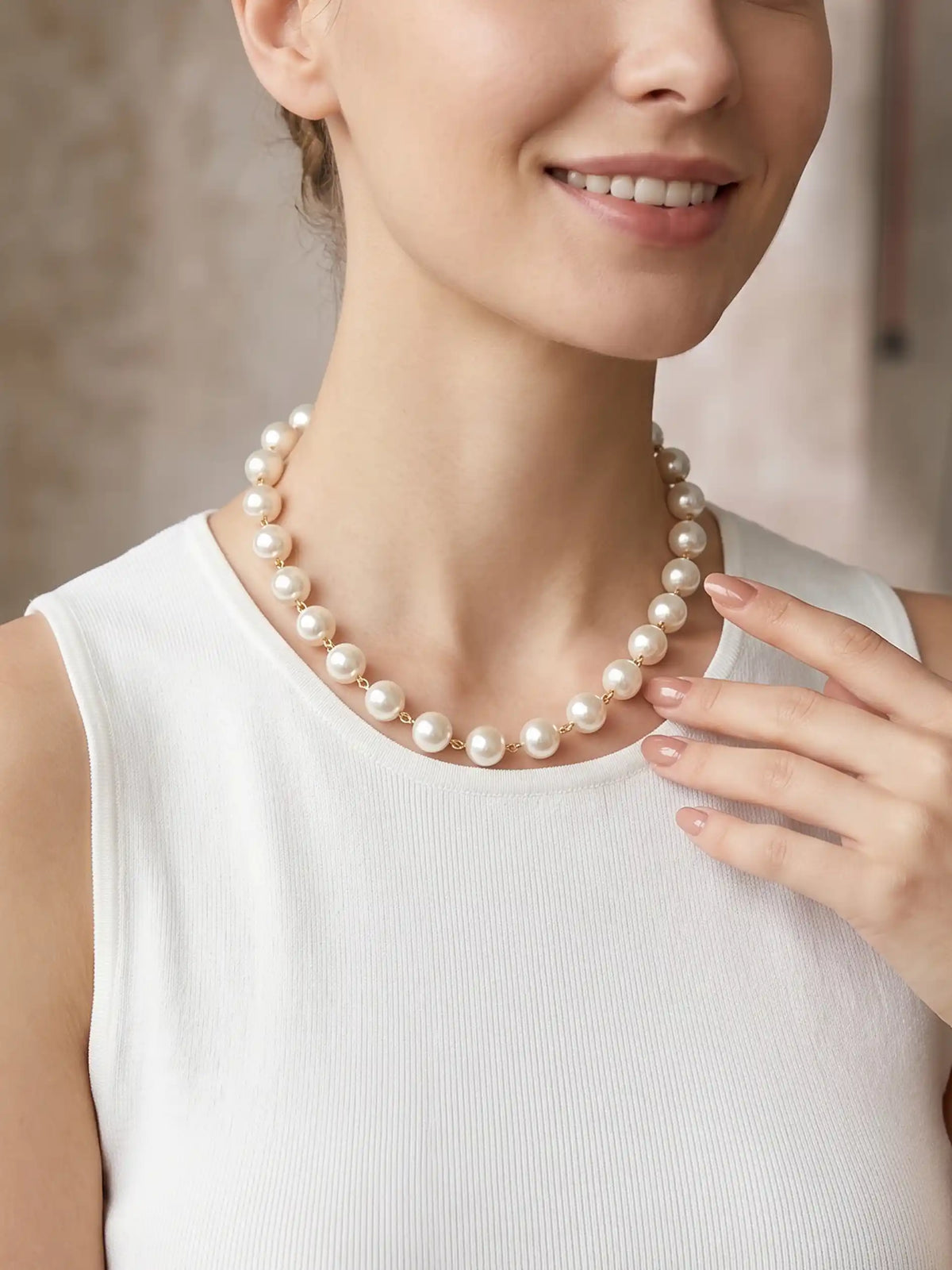 A medium close-up portrait of a smiling woman, looking directly at the camera. She is wearing a white sleeveless dress and a single-strand pearl necklace. The background is a slightly blurred studio environment with wood paneling.