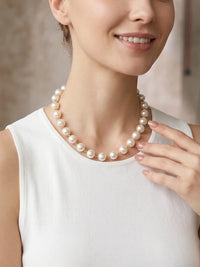 A medium close-up portrait of a smiling woman, looking directly at the camera. She is wearing a white sleeveless dress and a single-strand pearl necklace. The background is a slightly blurred studio environment with wood paneling.