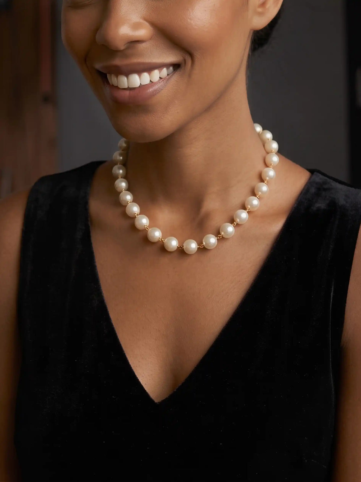 A medium close-up portrait of a smiling woman, looking directly at the camera. She is wearing a black sleeveless dress and a single-strand pearl necklace. The background is a slightly blurred studio environment with wood paneling.
