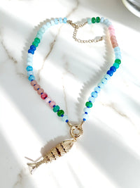 A beaded necklace with a gold fish pendant, laid out on a white marble surface with soft shadows. The necklace features light blue, green, and pink translucent beads with a gold clasp and fish charm.