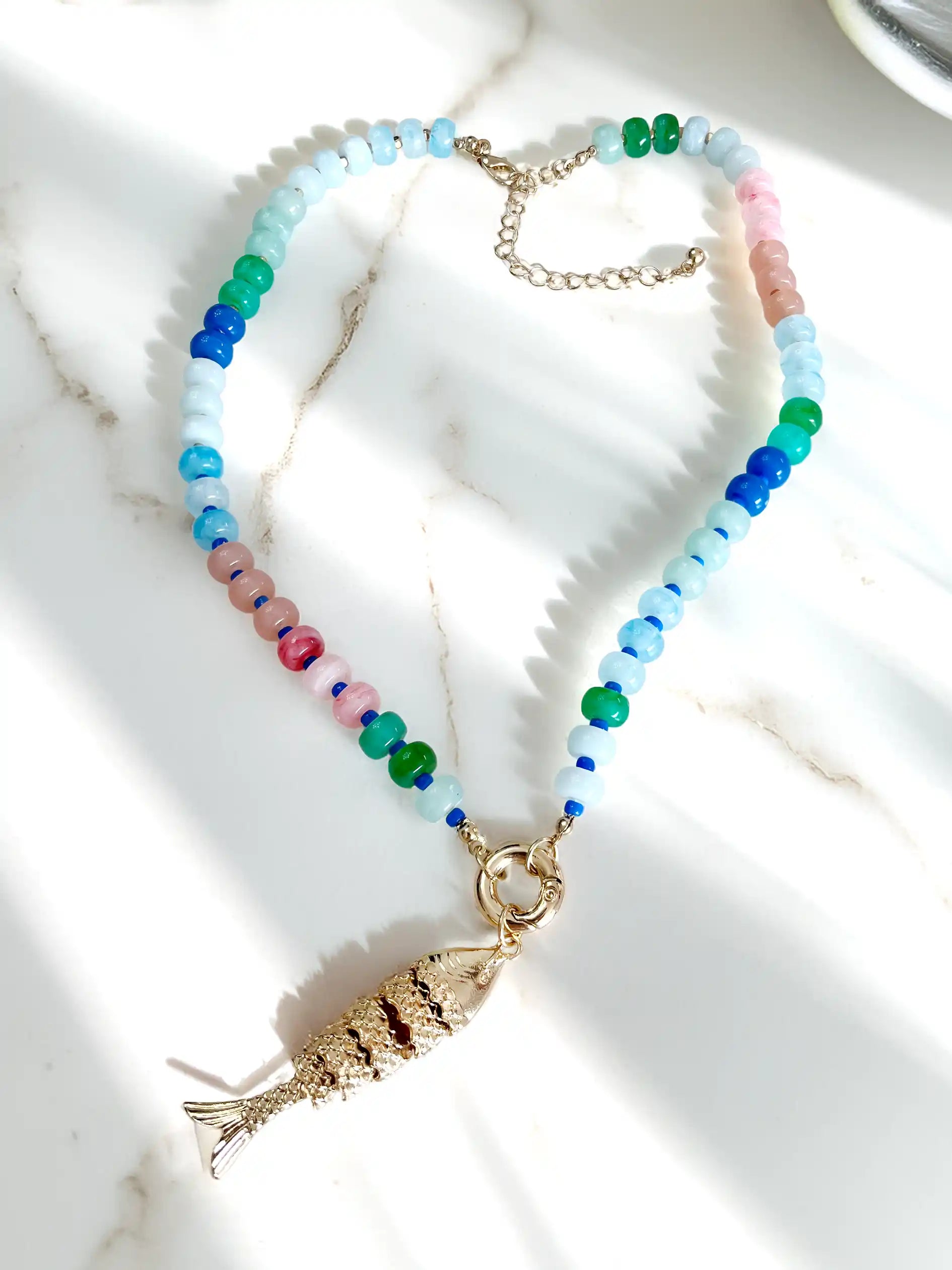 A beaded necklace with a gold fish pendant, laid out on a white marble surface with soft shadows. The necklace features light blue, green, and pink translucent beads with a gold clasp and fish charm.