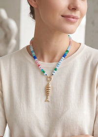 A close-up of a woman wearing a colorful beaded necklace with a gold fish-shaped pendant, paired with a cream-colored knit top.