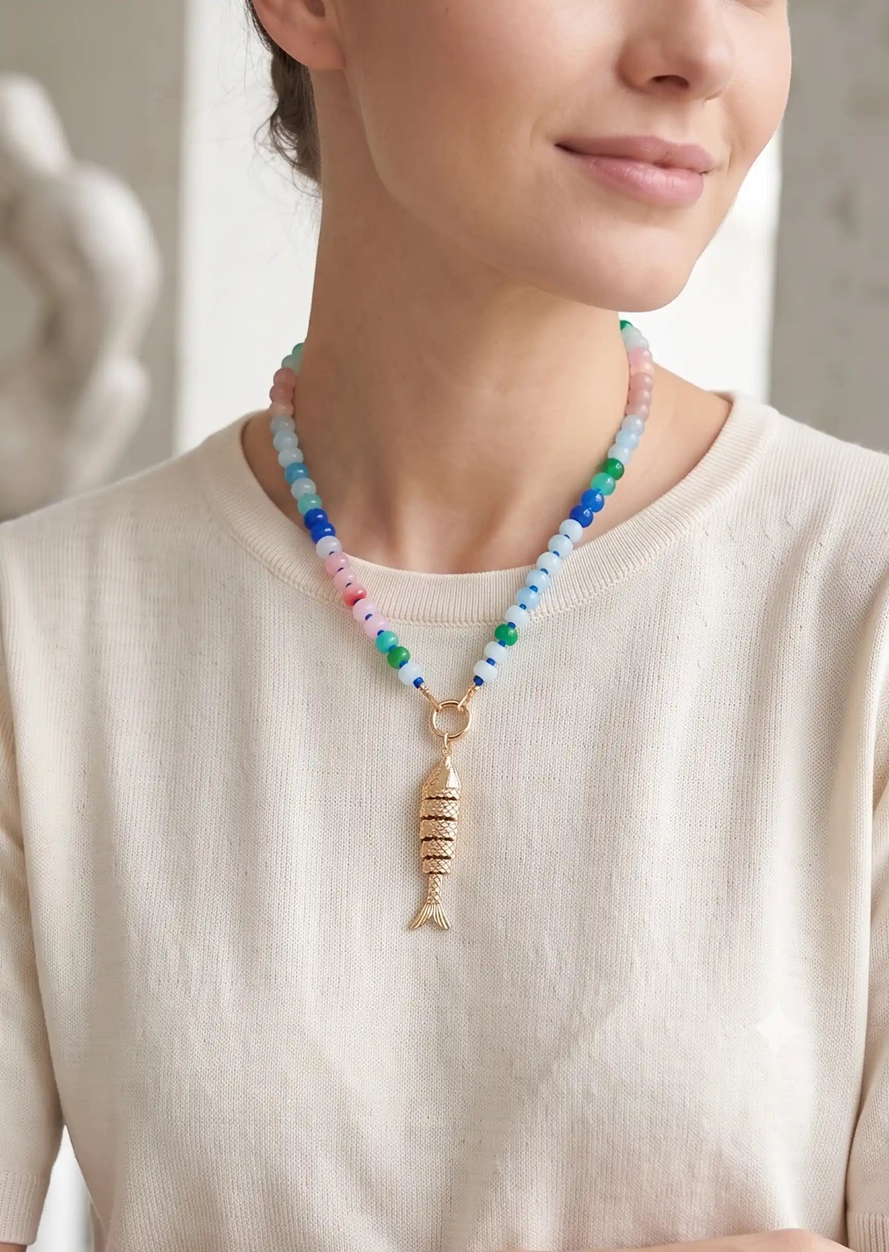 A close-up of a woman wearing a colorful beaded necklace with a gold fish-shaped pendant, paired with a cream-colored knit top.