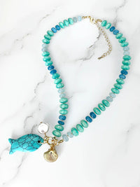 Turquoise and blue beaded statement necklace featuring glossy rondelle beads in aqua, teal, and deep blue tones. The necklace is accented with a large turquoise fish charm, a freshwater pearl, and a gold-tone scallop shell charm
