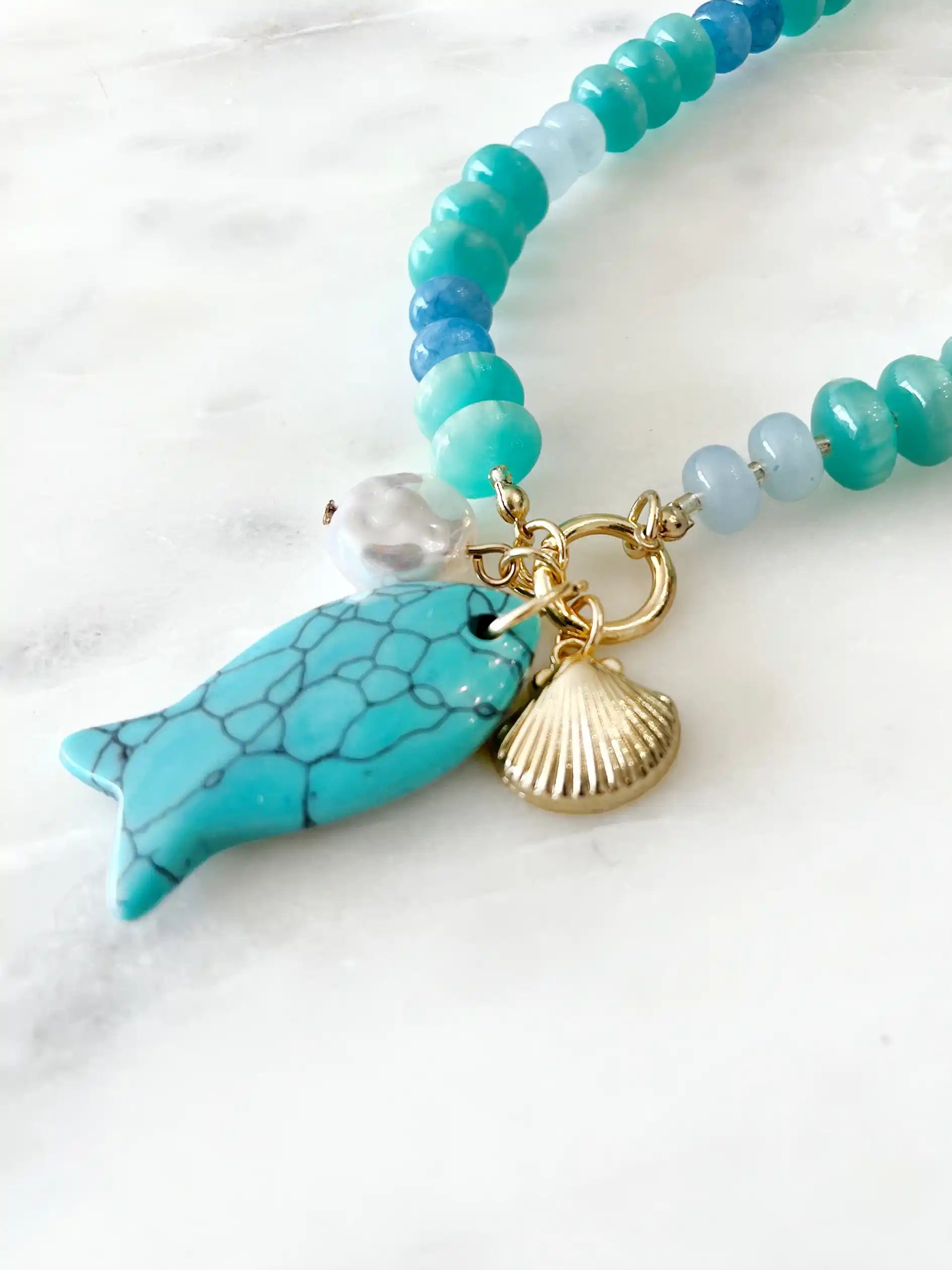 Turquoise and blue beaded statement necklace featuring glossy rondelle beads in aqua, teal, and deep blue tones. The necklace is accented with a large turquoise fish charm, a freshwater pearl, and a gold-tone scallop shell charm