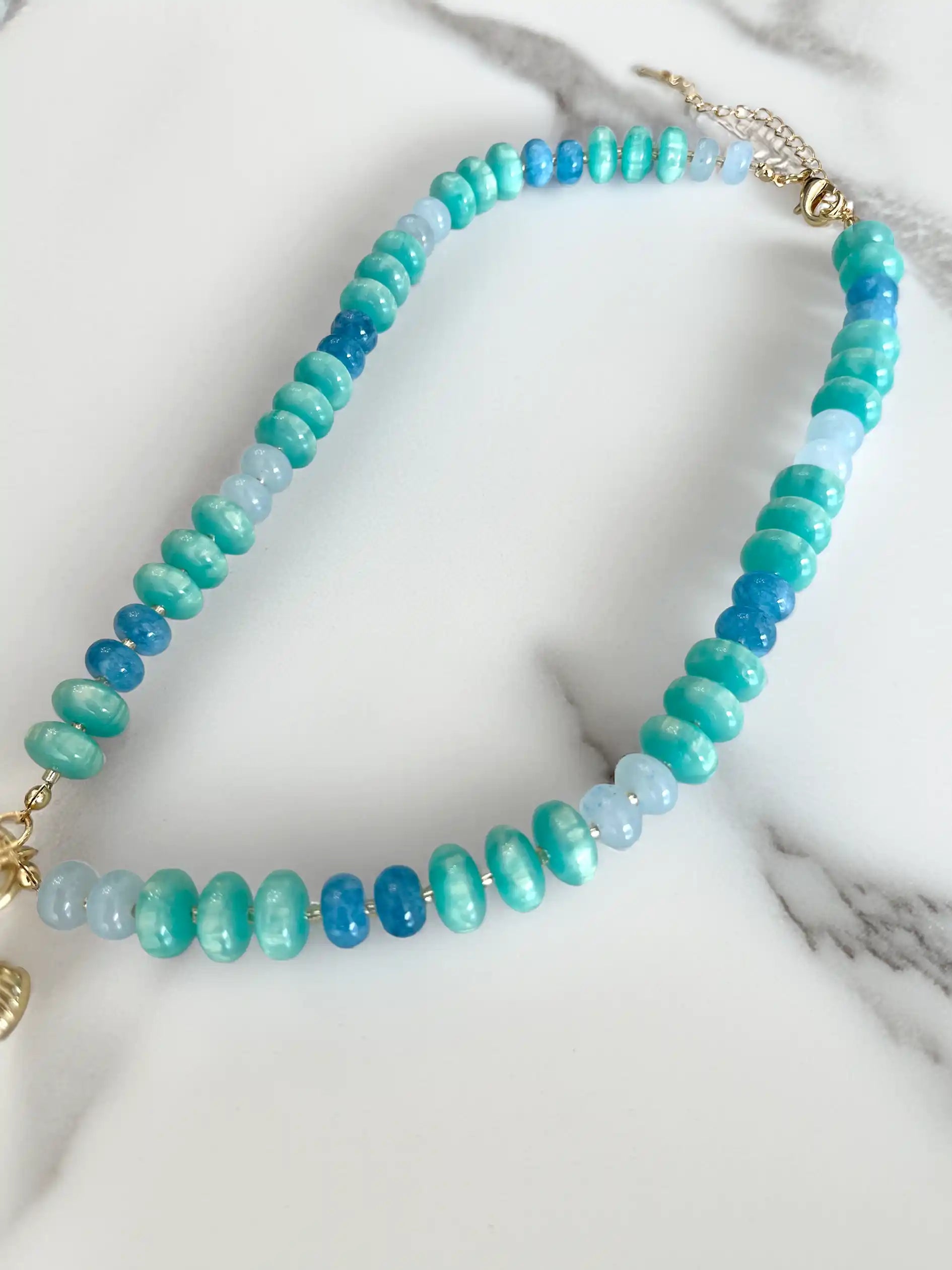 Turquoise and blue beaded statement necklace featuring glossy rondelle beads in aqua, teal, and deep blue tones. The necklace is accented with a large turquoise fish charm, a freshwater pearl, and a gold-tone scallop shell charm