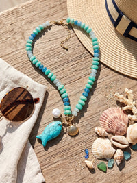 Turquoise and blue beaded statement necklace featuring glossy rondelle beads in aqua, teal, and deep blue tones. The necklace is accented with a large turquoise fish charm, a freshwater pearl, and a gold-tone scallop shell charm