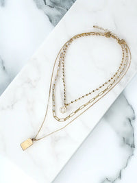 A gold-toned three-layer necklace against a plain white background. The top layer features a short beaded chain with a simple circular ring pendant. The middle layer is a wider chain with elongated oval links. The bottom, longest layer is a fine chain with a solid rectangular bar pendant.