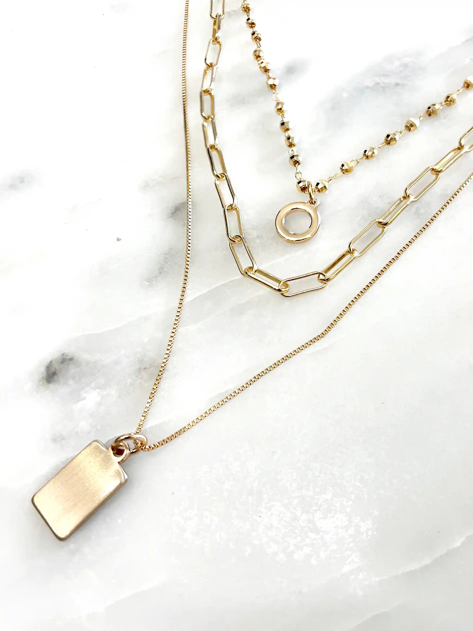 A gold-toned three-layer necklace against a plain white background. The top layer features a short beaded chain with a simple circular ring pendant. The middle layer is a wider chain with elongated oval links. The bottom, longest layer is a fine chain with a solid rectangular bar pendant.