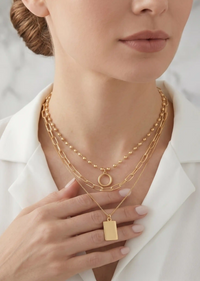 A close-up of a woman wearing layered gold necklaces, including a beaded chain, a paperclip chain with a ring pendant, and a longer chain with a rectangular tag pendant, paired with a white collared shirt.