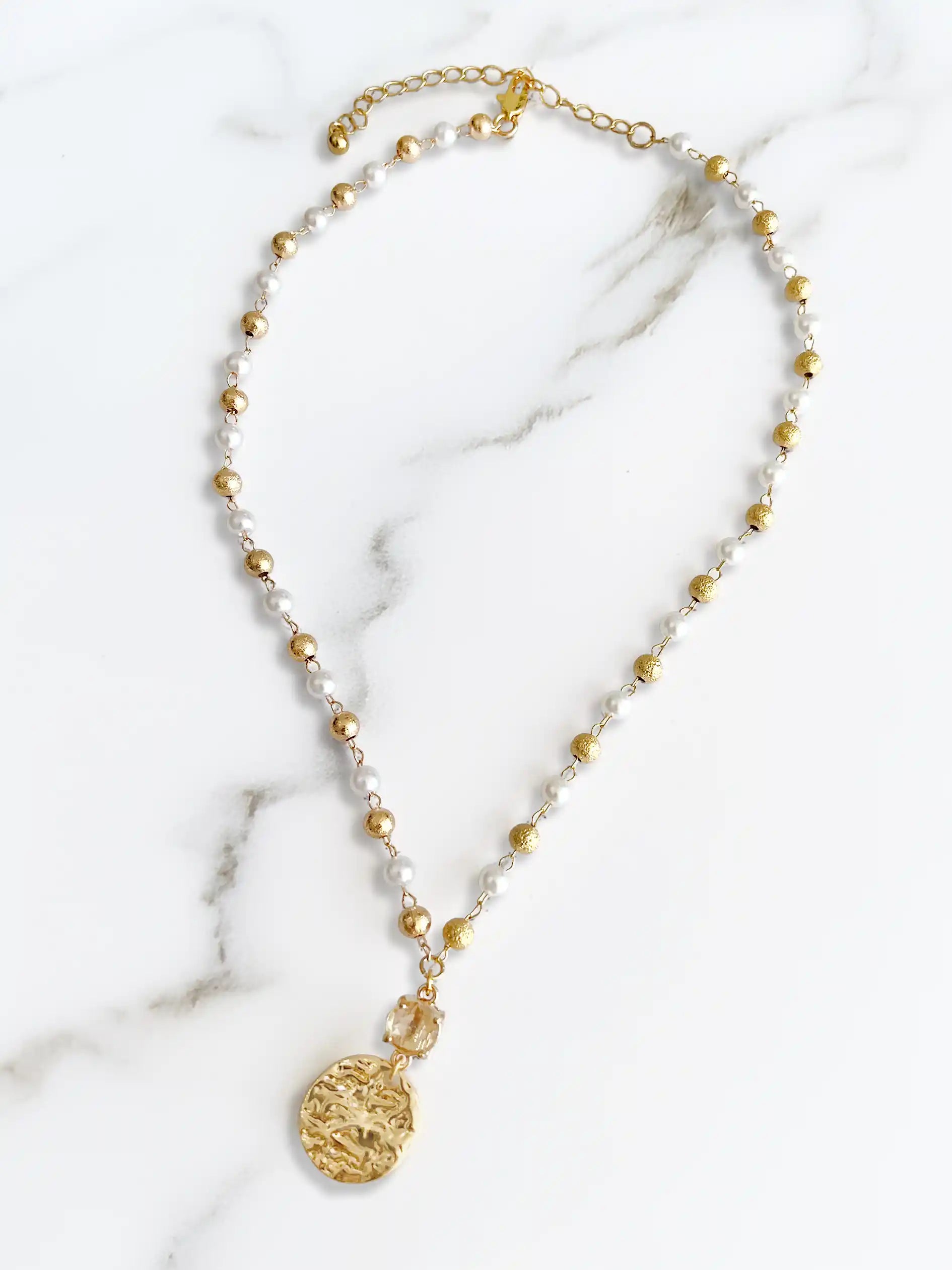 A delicate beaded necklace featuring alternating white pearls and small gold beads, with a textured gold coin pendant and matching gold chain extender, displayed on a white marble background.