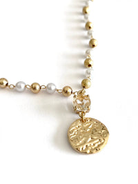 A close-up of a delicate necklace featuring gold-tone beads, white pearl-like beads, a clear faceted crystal charm, and a textured round gold coin pendant, set against a plain white background.