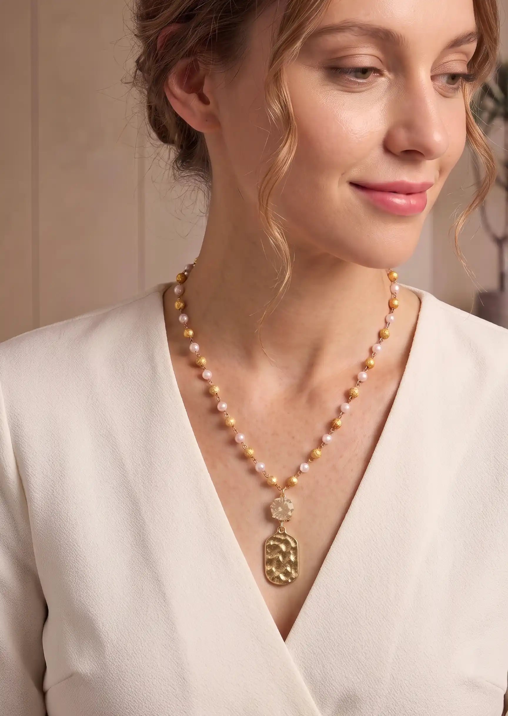 A woman wearing a delicate gold necklace with a large pendant and alternating gold and pale pink pearls, paired with a cream-colored blouse.