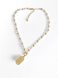 A delicate gold-tone beaded necklace with a textured, polished gold tag pendant, displayed on a clean white textured background.