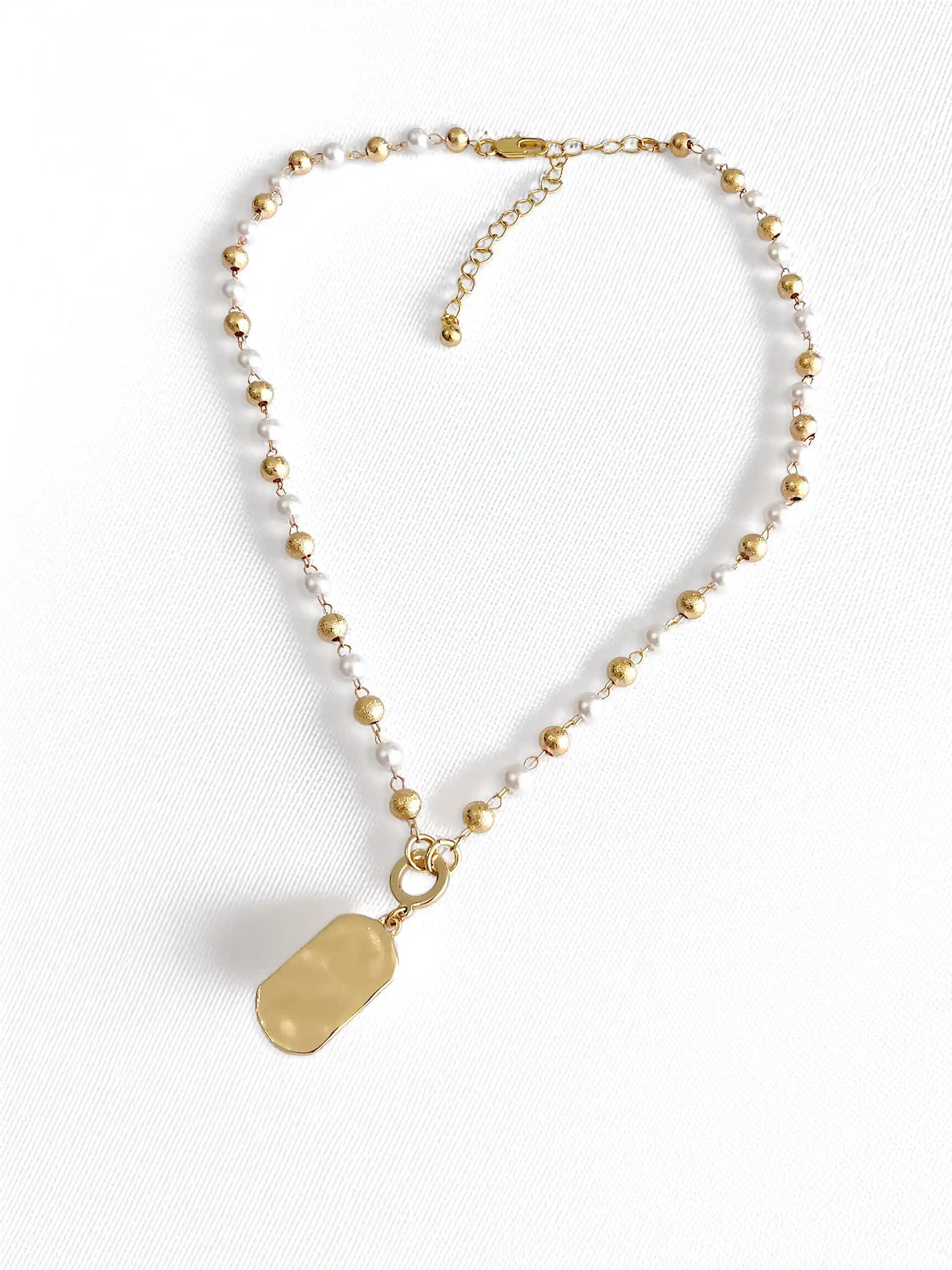A delicate gold-tone beaded necklace with a textured, polished gold tag pendant, displayed on a clean white textured background.