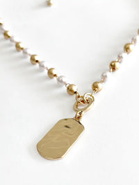 A gold-tone beaded necklace with a hammered rectangular dog tag pendant, displayed on a clean white background.