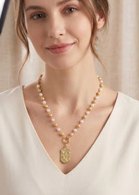 A woman wearing a delicate necklace with white pearls and gold-toned beads, featuring a textured gold pendant, photographed in a warm, softly lit setting.