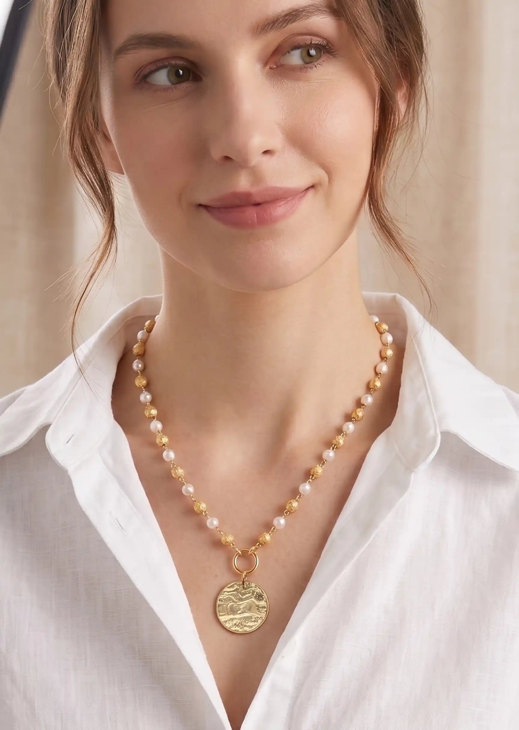 A close-up of a woman wearing a delicate necklace with white and gold pearls and a round gold pendant, paired with a white collared shirt.