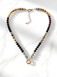 A semi-precious stone beaded necklace featuring black, white, tan, and reddish-brown beads, finished with a gold-tone round carabiner clasp and a small circular gold charm, displayed on a white marble surface.