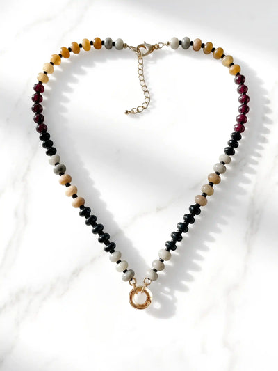 A semi-precious stone beaded necklace featuring black, white, tan, and reddish-brown beads, finished with a gold-tone round carabiner clasp and a small circular gold charm, displayed on a white marble surface.