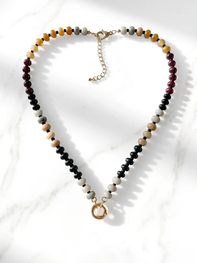 A semi-precious stone beaded necklace featuring black, white, tan, and reddish-brown beads, finished with a gold-tone round carabiner clasp and a small circular gold charm, displayed on a white marble surface.