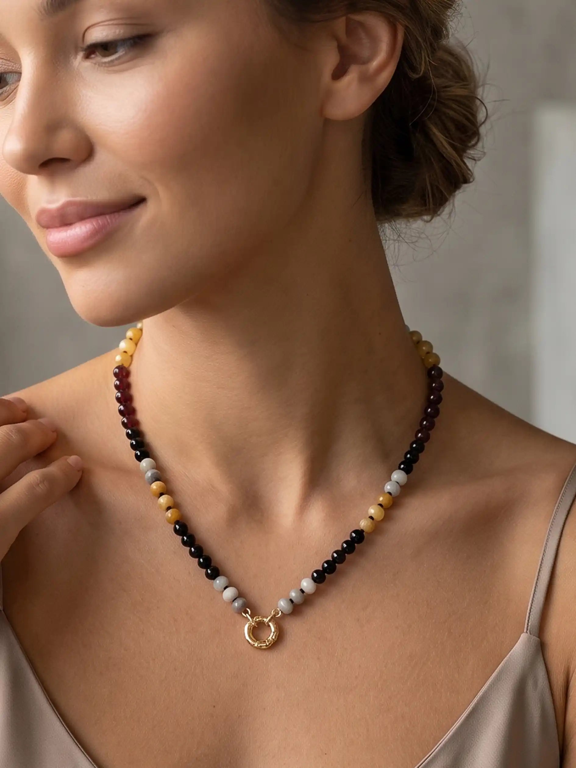 A woman wearing a semi-precious stone beaded necklace featuring black, white, tan, and reddish-brown beads, finished with a gold-tone round carabiner clasp and a small circular gold charm, displayed on a white marble surface.