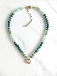 A semi-precious stone beaded necklace with faceted green and blue gradient beads and gold-tone round carabiner, displayed on a white marble surface.