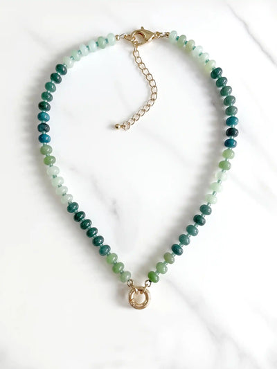 A semi-precious stone beaded necklace with faceted green and blue gradient beads and gold-tone round carabiner, displayed on a white marble surface.