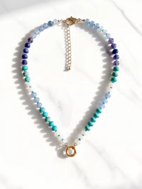 A semi-precious stone beaded necklace with faceted round beads transitioning through shades of blue, teal, green, and purple, accented by small white beads and gold-tone circle carabiner, displayed on a white marble surface.